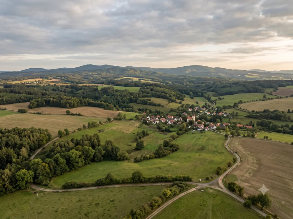 Letecký pohled na obec v českém pohraničí — kostel, pole a zalesněné kopce za soumraku